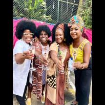 Quatro mulheres negras sorriem e brindam com taças de espumante em um ambiente ao ar livre e colorido. Estão vestidas com roupas vibrantes e estilosas, e usam acessórios marcantes, como brincos grandes, turbantes e óculos. O fundo rosa vibrante e a energia da imagem transmitem alegria, celebração e união.