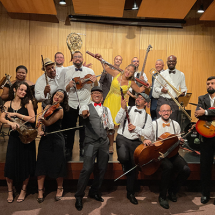 A imagem mostra um grupo musical posando de forma descontraída em um palco com parede de madeira e instrumentos ao fundo. São quatorze integrantes, todos sorridentes e vestidos elegantemente — alguns com trajes formais, como gravatas borboleta, e outros com roupas mais coloridas. Entre os instrumentos estão violinos, violoncelo, guitarra, contrabaixo, sopros e percussão. A atmosfera é alegre e celebrativa, transmitindo o entusiasmo coletivo pela música.