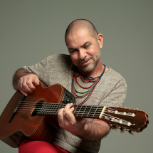 A imagem mostra o cantor e compositor paulistano Paulo Padilha tocando violão. Ele está sorrindo levemente, em uma pose descontraída, usando uma camiseta cinza, calça vermelha e colares coloridos. O fundo é neutro, destacando Padilha e seu instrumento. A cena transmite leveza e intimidade, refletindo o estilo bem-humorado e envolvente do artista.