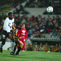 A imagem mostra um momento de intensa disputa durante uma partida entre Peñarol e Botafogo. Jogadores do Peñarol, com uniforme listrado em preto e amarelo, e do Botafogo, com uniforme branco e preto, acompanham atentos a trajetória da bola no ar. A cena se passa em um estádio com arquibancadas lotadas, transmitindo a energia vibrante de um jogo competitivo. Ao fundo, um garoto com roupa adidas vermelha observa a ação de perto.