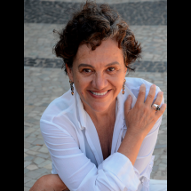 Adriana Vieira Lomar, romancista e contista, é uma mulher de pele clara e cabelos curtos, castanhos e cacheados. Ela sorri com simpatia, olhando diretamente para a câmera. Usa uma camisa branca com decote em V, brincos longos e um anel prateado com pedra escura na mão direita, apoiada sobre o ombro esquerdo. Ao fundo, há um chão de pedras portuguesas em tons claros e escuros.