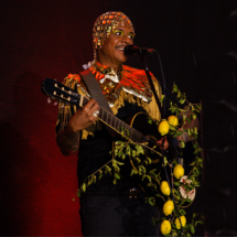Pessoa se apresenta no palco cantando e tocando violão, decorado com limões e ramos verdes. Usa uma roupa marcante com franjas douradas e um adorno brilhante na cabeça feito de miçangas coloridas. A expressão é de alegria e envolvimento com a performance. A iluminação destaca o artista contra um fundo escuro com luz vermelha, criando uma atmosfera vibrante e performática.
