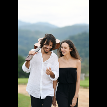 Os cantores e violonistas Antonia Medeiros e Guilherme Imia, integrantes do Duo Dois a Dois, caminham ao ar livre em meio à natureza, com montanhas ao fundo. Guilherme, de barba e cabelos ondulados, veste camisa branca e carrega um violão apoiado no ombro. Antonia, de cabelos cacheados e soltos, usa um macacão preto sem alças. Eles trocam sorrisos e parecem conversar, transmitindo leveza, sintonia e conexão artística.