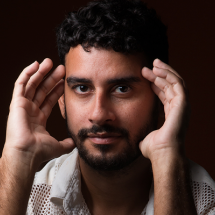 Homem de pele morena, cabelo cacheado curto e barba bem aparada, olha diretamente para a câmera com expressão serena. Ele está com as mãos levantadas próximas ao rosto, como se estivesse enquadrando ou refletindo. Usa uma camisa clara com textura vazada nos ombros. O fundo é escuro, destacando seu rosto iluminado.