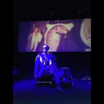 Na imagem, uma pessoa sentada em um banco no centro do palco é iluminada por uma luz azul intensa, que cria um efeito dramático. A figura veste um terno e usa óculos escuros, com postura relaxada e expressiva. Ao fundo, uma projeção com imagens difusas e simbólicas contribui para uma atmosfera cinematográfica e enigmática. A composição remete ao universo do teatro performático ou do cinema experimental, evocando mistério e intensidade visual.