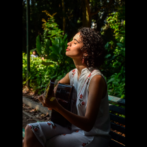 A cantora e violonista Katarina Assef aparece sentada em um banco de madeira em meio à vegetação exuberante, tocando violão. Com cabelos cacheados curtos, ela usa uma roupa clara com detalhes florais. De olhos fechados e rosto levemente inclinado para cima, parece absorver a música. A luz natural destaca seu perfil, criando uma atmosfera serena, sensível e contemplativa.