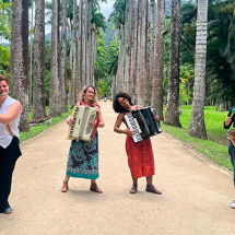 A imagem mostra as integrantes do grupo de forró Luz de Candeeiro em uma alameda cercada por palmeiras imperiais. Da esquerda para a direita, estão: Maria Souto tocando flauta, Marcela Coelho e Joana Araújo nas sanfonas, e Georgia Camara na percussão. As musicistas estão sorrindo e transmitindo alegria em meio ao cenário tropical, unindo natureza e cultura popular em uma atmosfera leve e vibrante.