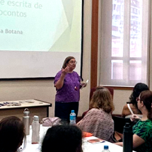 Imagem de oficina de escrita criativa em andamento, conduzida por Cecilia Botana, conforme indicado na projeção ao fundo com o título "Oficina de escrita de Microcontos". Cecilia, veste uma blusa roxa e está em pé diante de uma turma atenta, gesticulando enquanto fala. Os participantes estão sentados em mesas com cadernos e garrafas de água, em um ambiente iluminado por janelas grandes, que indicam tratar-se de uma sala em um prédio antigo, possivelmente histórico. A atmosfera é de concentração e interesse.