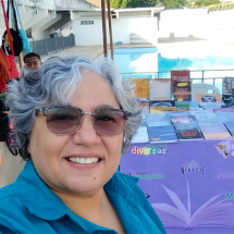 Mulher de cabelos grisalhos e curtos, usando óculos escuros e camisa azul, sorri para a câmera em uma selfie. É Léa Carvalho, publisher da Editora Metanoia, especializada em Gênero, Sexualidade e Diversidade. Ao fundo, há uma mesa com diversos livros expostos sobre uma toalha roxa com palavras como “diversas”, “Metanoia” e “Autor(a)Me”. Também há bolsas coloridas penduradas à esquerda e uma piscina ao fundo, sugerindo um evento ao ar livre, como uma feira cultural ou literária.