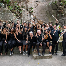 Um grupo grande de músicos posa ao ar livre diante de uma parede de pedra rodeada por vegetação. Todos estão vestidos de preto e exibem seus instrumentos com alegria, como flautas, trombones, violinos e clarinetes. Muitos sorriem, erguendo os instrumentos no ar em um gesto de celebração. A cena transmite um forte senso de união, entusiasmo e espírito coletivo em torno da música.