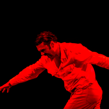 Homem em cena performática com iluminação totalmente vermelha, destacando seu corpo contra o fundo preto. Ele veste roupa social clara e está em movimento, com os braços estendidos e o tronco levemente inclinado para frente, transmitindo intensidade e expressão corporal. A imagem sugere um momento dramático ou coreográfico, com forte carga emocional e estética teatral.