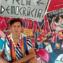 A imagem mostra o artista visual Alex Frechette sentado em frente a um mural vibrante que retrata uma cena de protesto, com cartazes e pessoas reunidas em manifestação. Ele veste uma camisa colorida com listras verticais e tem expressão séria. No fundo, lê-se frases como "Pela Democracia", "Ditadura Nunca Mais!" e "Esfacelaram o Evalantim!".
