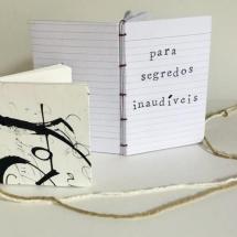A imagem apresenta dois cadernos artesanais sobre uma superfície branca. O caderno à direita está aberto, revelando páginas pautadas e a frase “para segredos inaudíveis” escrita em letra mecânica. Sua encadernação é feita com costura exposta em fio vermelho. O caderno à esquerda é menor, tem capa decorada com traços caligráficos abstratos em preto sobre fundo branco e está fechado. Um barbante cru e uma linha vermelha conectam os cadernos, sugerindo uma relação entre eles, remetendo a um conceito artístico.