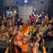 Pessoas de diferentes idades e estilos estão sentadas em um auditório pequeno, sorrindo e acenando para a câmera. Na frente, três pessoas se destacam, incluindo uma drag queen com peruca loira e figurino chamativo, que segura um microfone. O ambiente é acolhedor e descontraído, sugerindo um evento cultural ou artístico com forte senso de comunidade e celebração da diversidade.