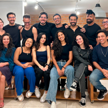 Grupo de 15 pessoas posa sorridente para a foto em ambiente interno com parede de tijolos e iluminação clara. O grupo é formado por homens e mulheres jovens, vestindo roupas casuais, majoritariamente em tons escuros. Sete pessoas estão sentadas à frente em bancos de madeira, enquanto as demais estão em pé atrás, com os braços sobre os ombros dos colegas. O clima é de amizade e descontração.
