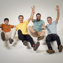 Os quatro integrantes do grupo vocal BeBossa posam sorridentes em estúdio com fundo branco, simulando estar em queda ou flutuação, com os braços erguidos e as pernas estendidas para a frente. Vestem camisetas coloridas (marrom, amarela, verde e azul) e calças casuais. A imagem transmite leveza, alegria e descontração, refletindo o espírito criativo e carismático do quarteto.