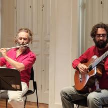 Os músicos Alexandre Bittencourt, na flauta, e Thomas Saboga, no violão, se apresentam em ambiente interno iluminado, sentados lado a lado diante de uma parede clara com portas antigas de madeira ao fundo. Ambos vestem camisas vermelhas. À esquerda, Alexandre toca flauta transversal com partitura à frente; à direita, Thomas dedilha o violão com expressão concentrada. A imagem transmite o clima intimista e refinado de uma apresentação de música de câmara.