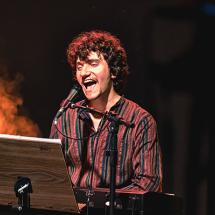 O cantor, compositor, regente e professor de música Gabriel Ruiz aparece em apresentação ao vivo, cantando com expressão vibrante enquanto toca teclado. Ele veste uma camisa listrada e está posicionado diante de um microfone, com uma estante de partituras à sua frente. Ao fundo, a iluminação cênica com fumaça alaranjada cria uma atmosfera intensa e envolvente, típica de uma performance musical.