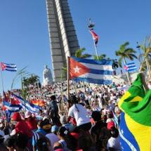 A imagem mostra uma multidão reunida em uma grande manifestação ou celebração pública em frente ao monumento a José Martí, na Praça da Revolução, em Havana, Cuba. Diversas pessoas carregam bandeiras de Cuba e também do Brasil, além de outros símbolos. O céu está limpo e azul, e palmeiras cercam o local, criando um cenário vibrante e ensolarado. A cena transmite um forte senso de união, engajamento e celebração coletiva.