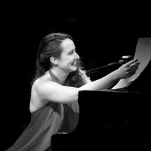 Imagem em preto e branco da cantora, compositora, pianista, atriz e neurocientista Julie Wein. Ela sorri enquanto está sentada ao piano, usando um vestido elegante. Com a mão direita, segura partituras, em um gesto que sugere preparação ou finalização de uma apresentação. Seu corpo está levemente inclinado para a frente, transmitindo leveza, carisma e envolvimento com a música.