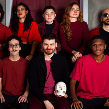 Os integrantes da banda carioca A Magnífica Máquina Maldita posam em grupo diante de uma parede cinza com portas vinho ao fundo. Quatro estão em pé na fileira de trás e quatro sentados à frente, todos vestindo roupas em tons de vermelho, bordô e preto. Um dos membros segura um crânio branco, evocando referências teatrais. Duas grandes máscaras expressivas estão apoiadas no chão à frente. A imagem transmite uma atmosfera artística, intensa e performática, característica da banda.