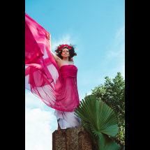 Mulher de braços erguidos veste vestido rosa vibrante e esvoaçante. Usa uma coroa de flores vermelhas e tem cabelos cacheados soltos. A imagem é captada de baixo para cima, com céu azul e árvores ao fundo, transmitindo sensação de liberdade, força e conexão com a natureza.