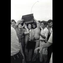 Fotografia em preto e branco de Juvenal Pereira, parte da exposição FotoRio 2025 no CCJF. A imagem retrata um momento de grande agitação popular: um homem de terno e gravata é cercado por várias pessoas, enquanto uma mala é erguida sobre sua cabeça para protegê-lo. A cena transmite a intensidade e o tumulto de um episódio coletivo marcante.