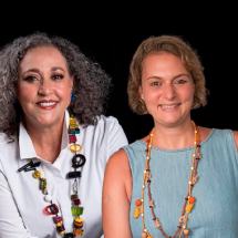 Retrato em fundo preto de duas mulheres sorrindo, lado a lado. À esquerda está Sandra Helena Pedroso, produtora e gestora cultural, com cabelos grisalhos cacheados na altura dos ombros. Ela veste camisa branca e um colar colorido com grandes contas. À direita está a jornalista Liana Varsano, de cabelos loiros curtos, usando blusa azul-clara sem mangas e colares em tons terrosos. Ambas transmitem simpatia e acolhimento.