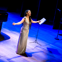 A soprano Chiara Santoro, mulher negra, apresenta-se no palco vestindo um longo vestido prateado. Ela canta de braços abertos e expressão intensa, transmitindo emoção. Ao fundo, aparecem parte de um piano de cauda e um pedestal com partitura. A cena é iluminada por luzes azuis, realçando a atmosfera solene da performance.