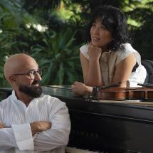 Fotografia do Duo Burajiru, formado por Fernando Thebaldi (violão) e Yuka Shimizu (piano). Em ambiente ao ar livre, Yuka está deitada sobre o piano com expressão atenta e relaxada, enquanto Fernando, sentado ao instrumento, cruza os braços e a observa com leve sorriso. Um violino repousa sobre o piano. Ao fundo, vegetação tropical completa a cena.