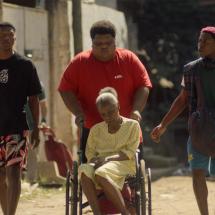 Cena do filme Kasa Branca. A imagem mostra três jovens caminhando por uma rua de terra enquanto um deles empurra uma senhora idosa em cadeira de rodas. O grupo transmite união e cuidado em meio ao ambiente simples da comunidade, refletindo afeto e solidariedade.