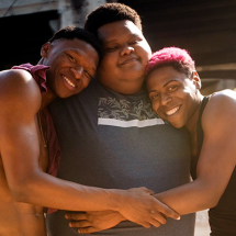Três jovens negros posam sorridentes em um abraço coletivo, transmitindo afeto e amizade. O rapaz ao centro veste camiseta cinza estampada, enquanto os dois ao lado, um com o cabelo descolorido em tom rosado e outro com o peito nu, encostam a cabeça em seus ombros. A cena, iluminada pela luz natural, destaca a conexão e a alegria compartilhada entre eles.
