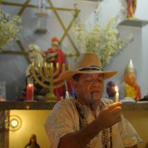 Homem com chapéu de palha e colares fuma cachimbo e aparece em frente a um altar religioso, segurando uma vela acesa com expressão de concentração e respeito. O altar é decorado com flores, imagens de santos e entidades, velas coloridas e um candelabro dourado, compondo um ambiente espiritual e ritualístico.