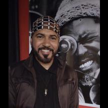 O pedagogo, curador, cineasta, roteirista e produtor Alek Lean sorri para a câmera, usando camisa preta, jaqueta marrom e um turbante estampado em marrom e branco. De barba bem aparada e expressão acolhedora, ele posa diante de um painel em preto e branco que mostra, em detalhe, o rosto de um homem sorridente, criando um cenário de forte atmosfera artística.