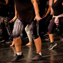 Grupo de pessoas pratica passos de dança charme em sala de ensaio. Vestindo roupas esportivas, como camisetas, leggings e tênis, algumas também estão descalças. A cena destaca o movimento das pernas em sincronia, transmitindo o ritmo marcado, o balanço característico e a energia coletiva dessa expressão cultural.