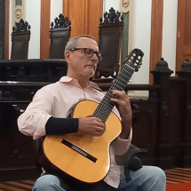 Foto da Sala de Sessões do CCJF. Ao centro, no tapete vermelho, sentado em uma cadeira, o violonista Nicolas de Souza Barros. Homem branco, cabelos grisalhos cortados rente à cabeça, óculos retangulares de hastes escuras. Ele usa uma camisa social clara, arregaçada até quase o cotovelo e calça jeans clara.. Em seu antebraço direito, ele usa uma munhequeira preta. Ele toca o violão apoiado em sua perna direita.