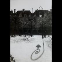 Foto em preto e branco de uma praça vazia em dia nublado, com clima que sugere frio. Árvores sem folhas projetam sombras intricadas no chão claro — que pode ser de pedra clara ou coberto por uma fina camada de neve. Um poste de luz com design curvo aparece à direita e sua sombra espiralada ocupa o primeiro plano. Ao fundo, há construções antigas com telhados inclinados e janelas pequenas, evocando arquitetura europeia. A cena transmite silêncio e contemplação.