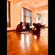 Três pessoas estão sentadas no chão de uma sala iluminada, com cortinas coloridas nas janelas ao fundo. Elas se reúnem em círculo sobre tapetes, em postura concentrada e introspectiva, sugerindo um momento de partilha, meditação ou prática ritual. O ambiente tem clima acolhedor, com sofá e almofadas ao lado, reforçando a sensação de intimidade e conexão.