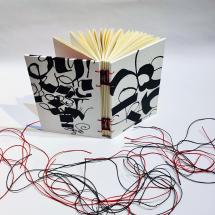 A imagem mostra um caderno artesanal aberto, com costura exposta em fios vermelhos e pretos. A capa é ilustrada com traços abstratos em preto sobre fundo branco, lembrando caligrafias e movimentos livres. Ao redor do caderno, sob uma superfície clara, estão espalhados fios soltos, reforçando o caráter manual e artístico da encadernação.
