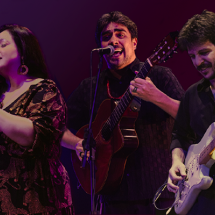 A imagem mostra os músicos Luiza Borges (voz), Thiago Thiago de Mello (voz e violão) e Diogo Sili (guitarras e arranjos) em apresentação. Luiza canta com expressão serena, Thiago interpreta com intensidade ao violão e Diogo toca guitarra com concentração. O fundo em tons de roxo e vermelho valoriza a cena, destacando a energia e harmonia do trio no palco.