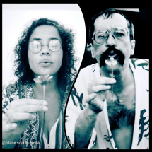 A imagem em preto e branco mostra Nilane Soares Costa e Paulo Leminski, ambos segurando uma flor próxima ao rosto e assoprando suas pétalas. Usando óculos redondos, encaram a câmera de forma expressiva, em um gesto poético que transmite delicadeza e a efemeridade do instante.