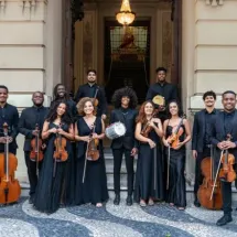 Integrantes da Camerata Jovem do Rio de Janeiro posam sorridentes em frente ao prédio histórico do Centro Cultural Justiça Federal (CCJF). O grupo, formado por 14 músicos vestidos com trajes formais pretos, segura instrumentos como violinos, violoncelos, contrabaixo, pandeiros e triângulo. A escadaria e o vitral iluminado ao fundo compõem o cenário elegante e acolhedor da foto.