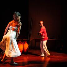 Cena em um palco iluminado por luz âmbar mostra a pianista Verónica Fernandes e o percussionista Anderson Vilmar em um momento de dança e expressão corporal. Verónica, de saia e top brancos, movimenta-se em diálogo com Anderson, que veste camiseta vermelha e calça branca. Ao fundo, um tambor reforça a atmosfera rítmica e ancestral da cena, marcada por energia, conexão e celebração da cultura afro-brasileira.
