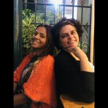 As escritoras Luciana Belém e Ednéia Rodrigues posam sorridentes, encostadas uma na outra, diante de um fundo com plantas. Luciana, à esquerda, veste um casaco alaranjado e tem cabelos longos; Ednéia, à direita, usa um casaco preto e apoia o queixo na mão, com expressão acolhedora. A imagem transmite leveza e cumplicidade entre as autoras.