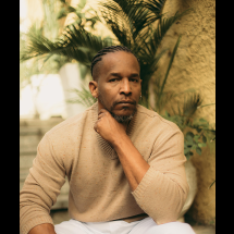 O cantor Alcides Sodré aparece sentado em um ambiente externo, cercado por folhagens. Ele olha diretamente para a câmera com expressão serena e introspectiva, apoiando o queixo sobre a mão. Usa uma blusa de tricô bege, e a iluminação suave valoriza seus traços e o clima de contemplação da cena.