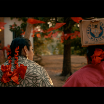 Cena do filme La Baláhna. Duas pessoas caminham por um espaço ao ar livre enfeitado com bandeirolas vermelhas entre as árvores. Vistas de costas, uma usa uma manta estampada e tranças adornadas com grandes laços vermelhos; a outra veste tecido vermelho e equilibra na cabeça uma caixa com o selo “Producto de México”. A luz dourada e o cenário evocam um clima de tradição e celebração popular.