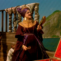 Cena do filme Carlota Joaquina, Princesa do Brazil, em que a atriz Marieta Severo interpreta a personagem principal. Ela aparece em um navio, vestida com traje luxuoso em tons de roxo e dourado, usando toucado e colares ornamentados. À sua frente, há um baú aberto, e ao fundo, o mar e o Pão de Açúcar compõem o cenário histórico da cena.