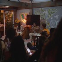 A imagem mostra um espaço cultural intimista, com pessoas sentadas assistindo a uma roda de conversa ou apresentação. À frente, duas pessoas estão no palco: uma mulher de cabelos longos e um homem falando ao microfone. O ambiente é decorado com murais artísticos na parede, iluminação aconchegante e plantas, criando uma atmosfera acolhedora e descontraída.