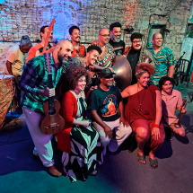 Integrantes da Orquestra Balogun posam juntos em um ambiente rústico, com paredes de pedra e iluminação colorida em tons de azul e vermelho. O grupo reúne músicos e artistas com instrumentos como violão, atabaque e outros tambores, formando uma composição vibrante. Todos sorriem e se aproximam para a foto, transmitindo união, celebração e a força da musicalidade afro-brasileira que marca o trabalho da orquestra.