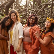A equipe responsável pelo Raízes – Festival de Arte Sustentável posa unida diante de um cenário natural formado por raízes e galhos entrelaçados. As quatro mulheres, vestidas em tons terrosos que dialogam com a proposta ecológica do festival, sorriem com acolhimento e cumplicidade. A composição transmite conexão com a natureza, ancestralidade e o espírito colaborativo que move o projeto.